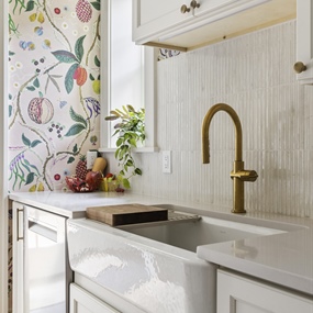 White farmhouse-style kitchen sink with brass faucet, subway tile backsplash, and floral wallpaper.