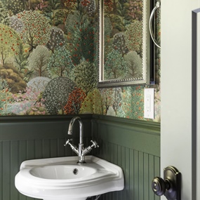 Close-up of a powder room remodel with a white pedestal sink, brass faucet, and dark green botanical wallpaper.